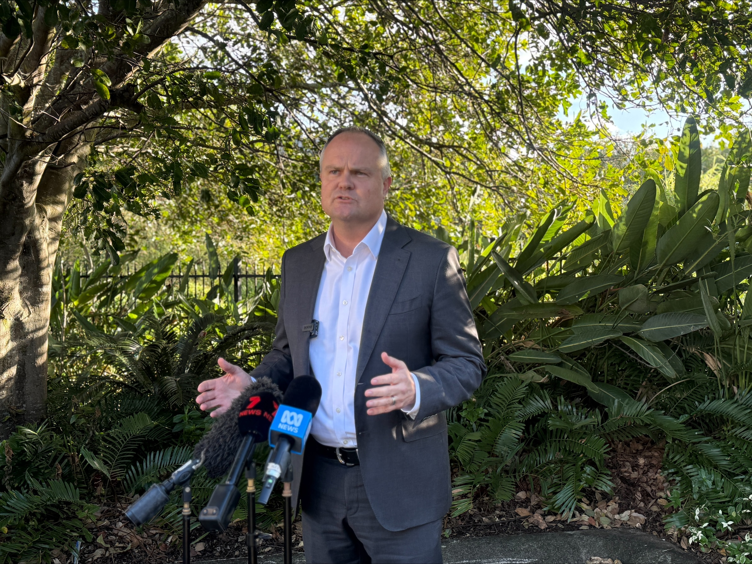 A man in a suit, speaking in front of media microphones, with trees and foliage in the background.