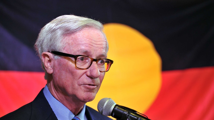 An older, bespectacled man with neatly combed silver hair, dressed in a dark suit, speaks in front of an Aboriginal flag.