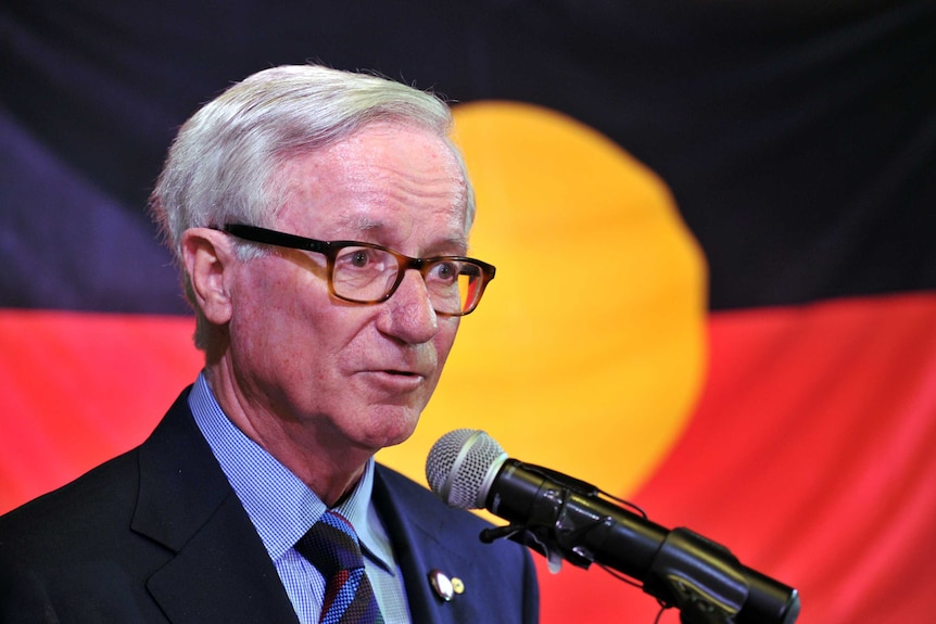 An older, bespectacled man with neatly combed silver hair, dressed in a dark suit, speaks in front of an Aboriginal flag.