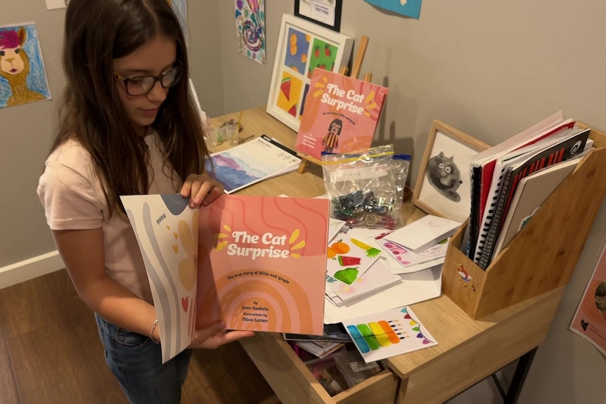 A girl with brown hair and glasses holds open a pink and orange picture book by a desk with art supplies on it. 