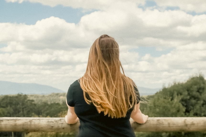 Foster care survivor Amy looks over a paddock with her back to the camera and the wind blowing in her hair.