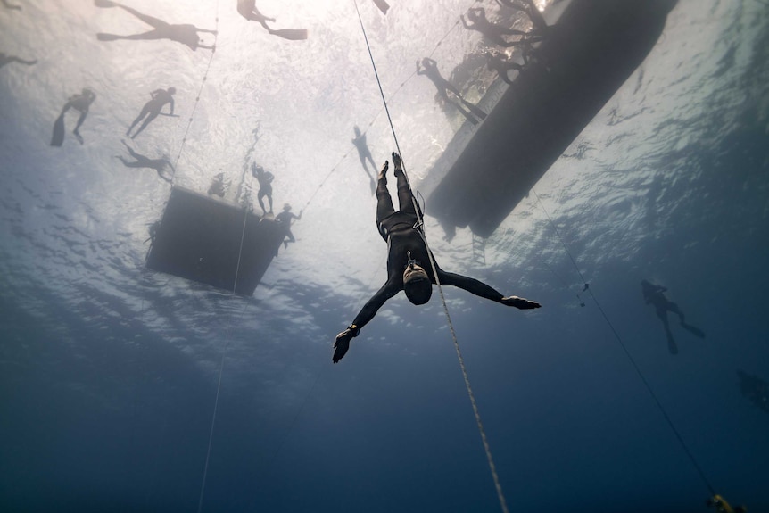 A woman in a wetsuit tethered to a guide line under the ocean with the silhouettes of other divers and boats in the background.