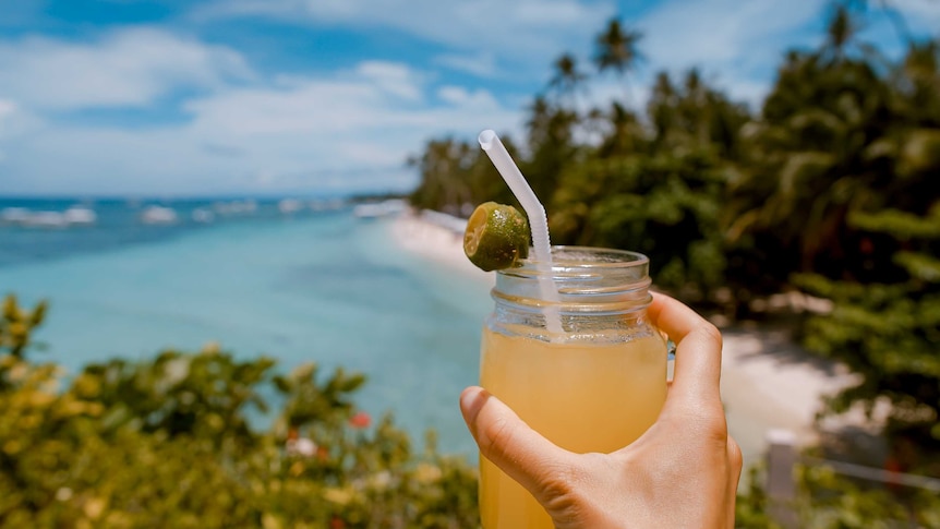 A hand holds a iced juice drink in a mason jar in front of a view of a palm tree-lined beach.