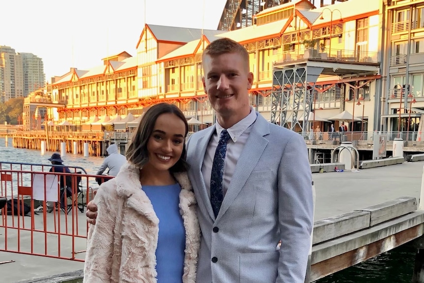 Hannah and her partner standing near a marina, with a building in the background and the Sydney Harbour Bridge just visible