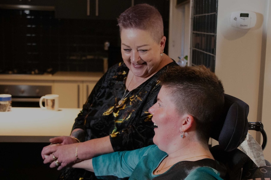 Lyn holds hands and smiles at her daughter Sarah, who sits beside her in a wheelchair.