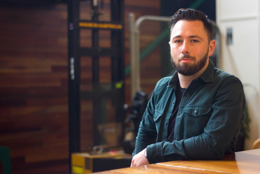 Bearded man with green shirt sits in wooden warehouse type room.