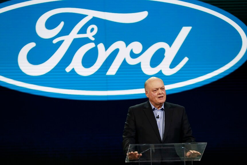 A man in a suit speaks from behind a clear lectern while standing in front of a large Ford logo