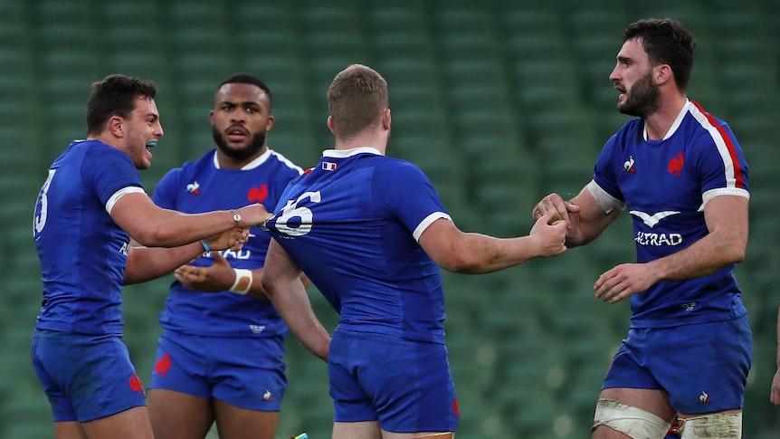 Four players wearing blue shirts grab at each other against a backdrop of green seats