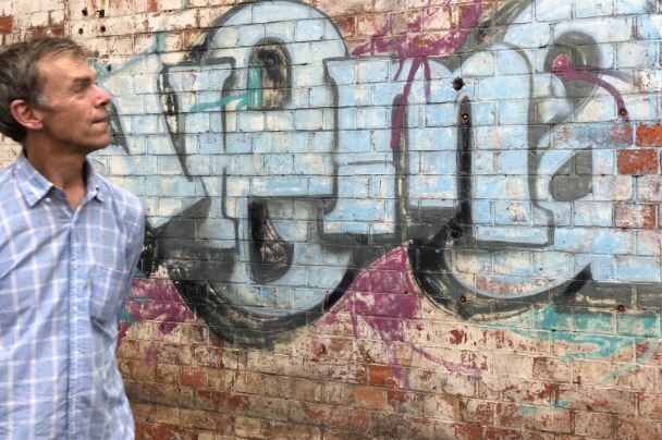 Bruce Jeffery walks in front of a brick wall with graffiti on it. He wears a blue shirt and tan trousers, and looks right.