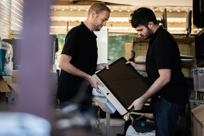Two men hold a big computer in a carport filled with electronics.