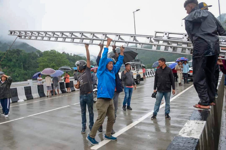 Two men help others lift a huge metal ladder as locals gather in the rain on a bridge.