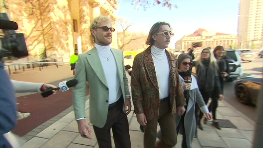 A man wearing a suit jacket and a man wearing a leather jacket and sunglasses outside a stone building