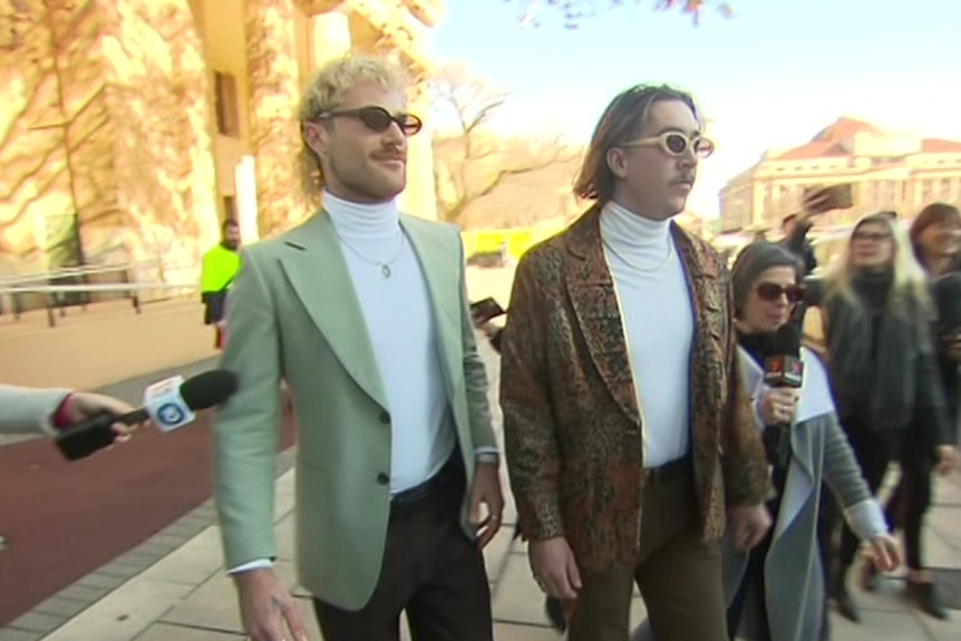 A man wearing a suit jacket and a man wearing a leather jacket and sunglasses outside a stone building