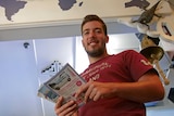 A young, dark-haired and bearded man smiles down at the camera holding a sheaf of tourist brochures.