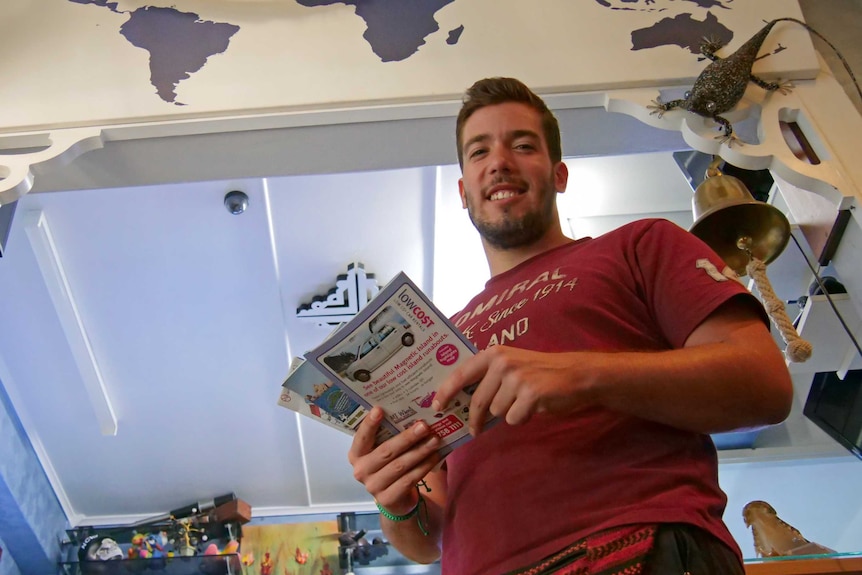 A young, dark-haired and bearded man smiles down at the camera holding a sheaf of tourist brochures.