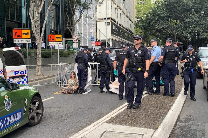 A young woman wearing camouflage sits chained by the neck to fencing, surrounded by police.