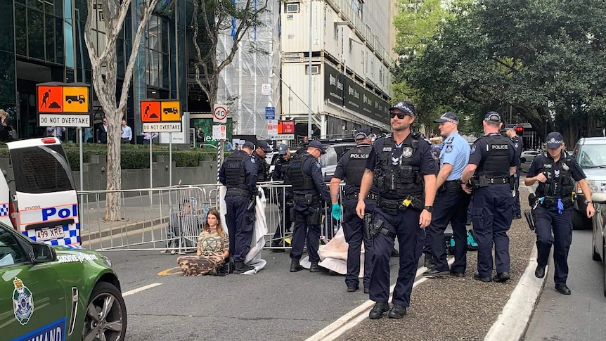 A young woman wearing camouflage sits chained by the neck to fencing, surrounded by police.