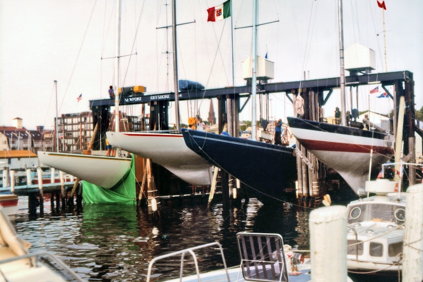 Four racing yachts lined up in a docking area. One of them has a tarpaulin covering its keel.