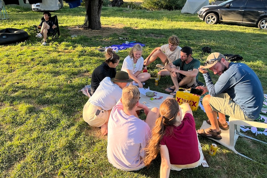 A group of young people sit on a rug playing a game at a pleasant campsite.