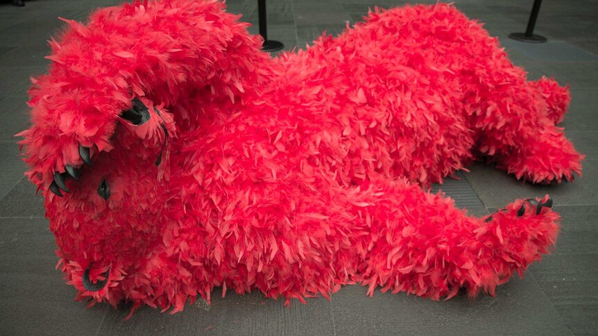 A neon red feathered bear lays on his back on a concrete tiled floor.