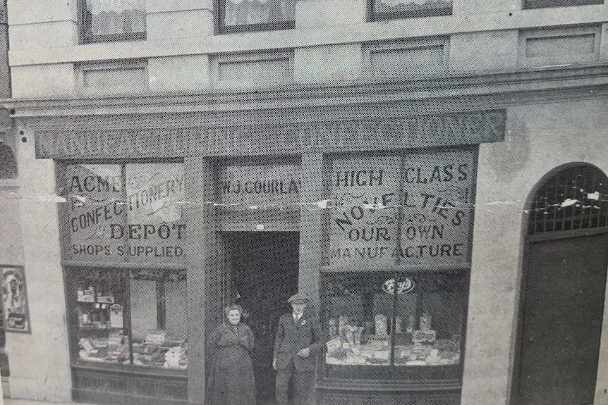 A black and white photo of an old confectionary store with two people standing at the entrance.