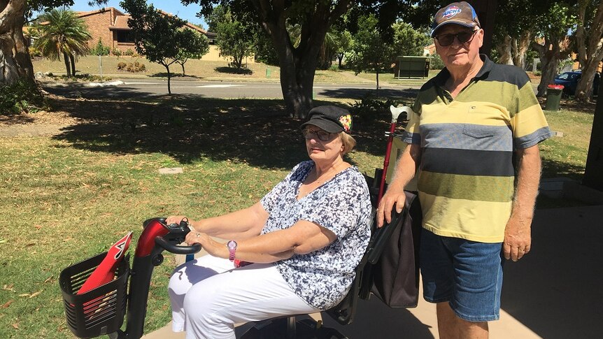 An older woman sits in a mobility scooter and a man stands behind her