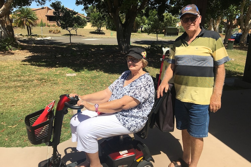 An older woman sits in a mobility scooter and a man stands behind her