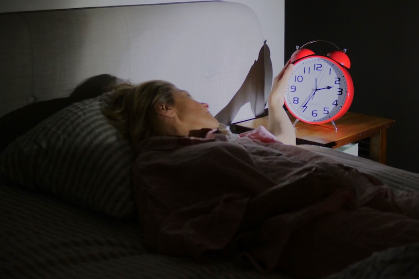 A woman lies awake in a bed looking at a big red clock