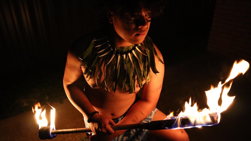 A man in traditional dress stands in darkness illuminated by large flames on either end of a long club.