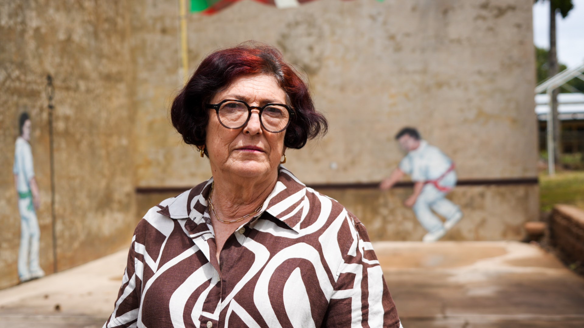 Marie-V Wherry standing in front of a concrete wall. She wears patterned clothes and has dark hair with red highlights.