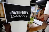 A sign saying #save the Daily Cambodia is stuck on a post as journalists work in the background.