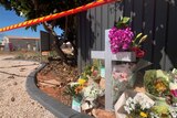 Image of a memorial set up beside a gravel road, a cross, flowers, bouquets under a blue sky and a tree.