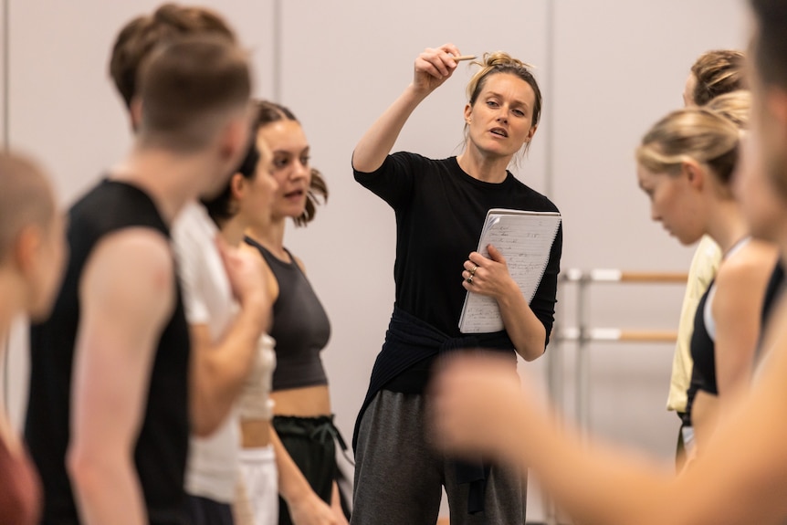 Stephanie Lake stands among a group of dancers in a dance studio holding a notepad in one hand and gesturing with the other.