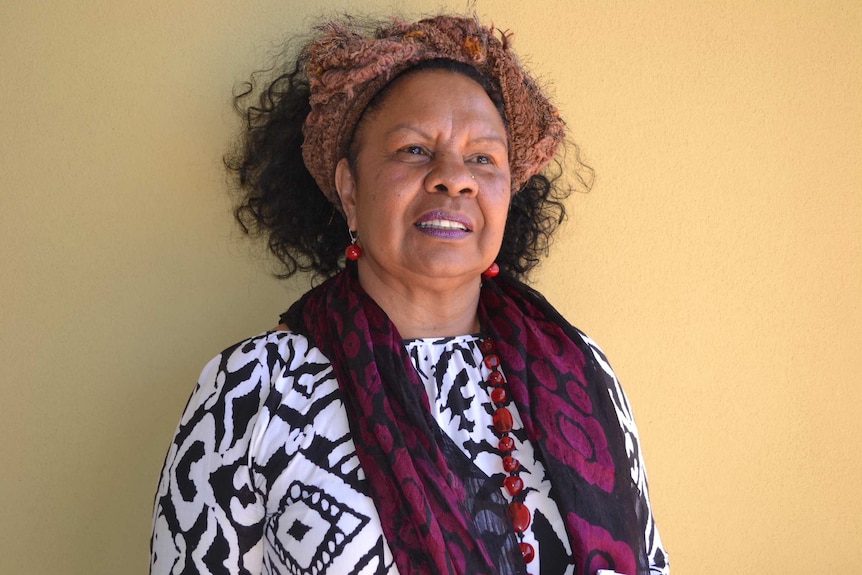 A mid shot of Nyikina traditional owner Anne Poelina standing against a wall wearing a white and black top and purple scarf.