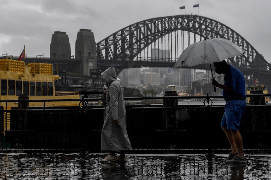 Person carrying an umbrella and person in a raincoat walk past the Sydney Harbour Bridge.