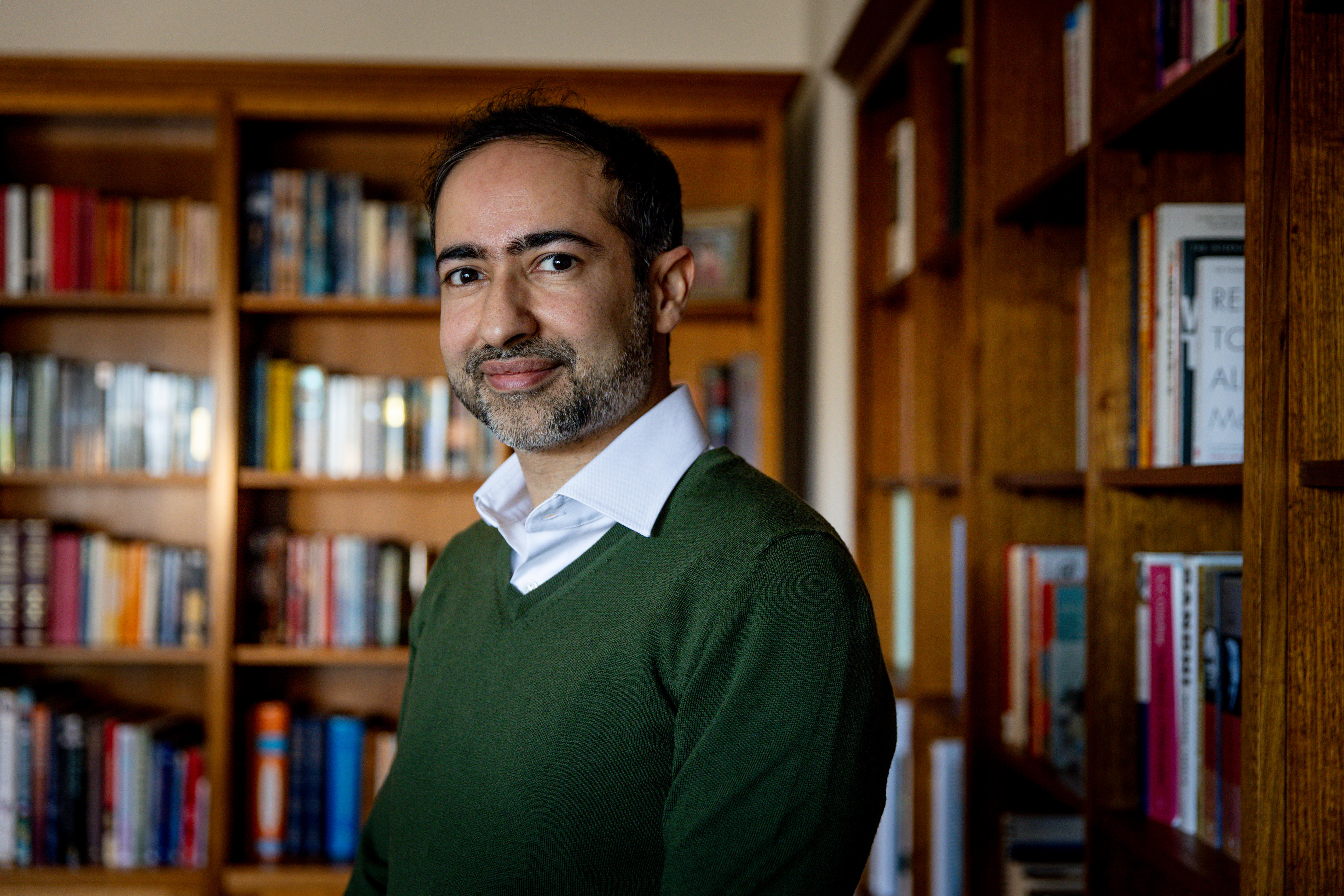 Rahul Khanna, dressed in a button-up shirt and green jumper, stands in front of bookshelves in an office.