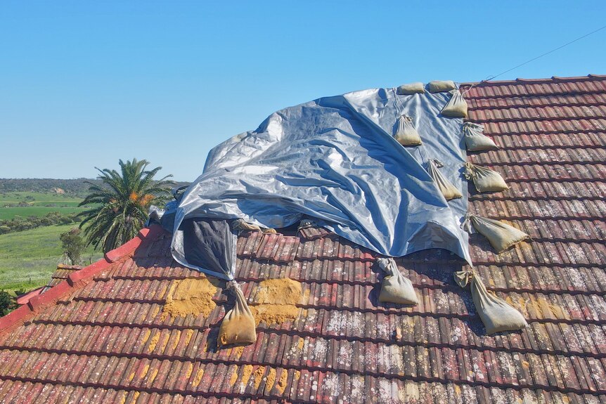 A silver tarpaulin on the edge of a pitched tiled roof is held down at the sides by sandbags. 