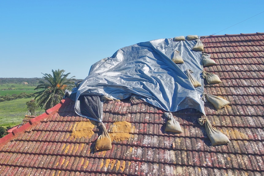 A silver tarpaulin on the edge of a pitched tiled roof is held down at the sides by sandbags. 