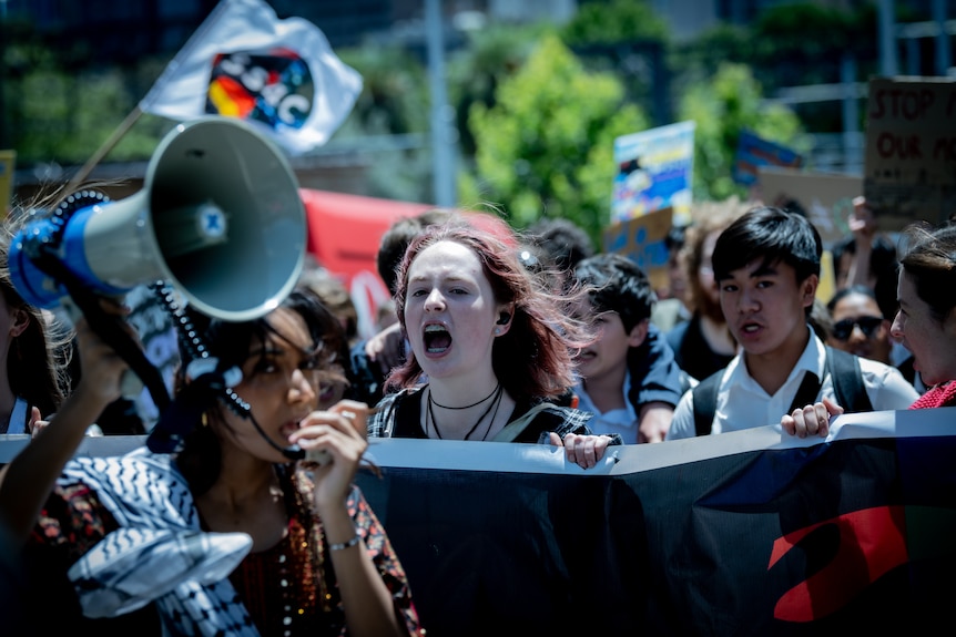 A girl in foreground with megaphone, behind a girl with braces holds a sign and shouts, more students march behind.