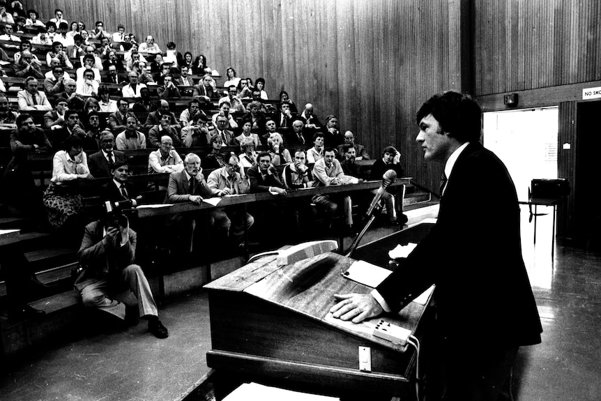 A middle-aged man in a suit stands behind a lectern and addresses a room full of academics.