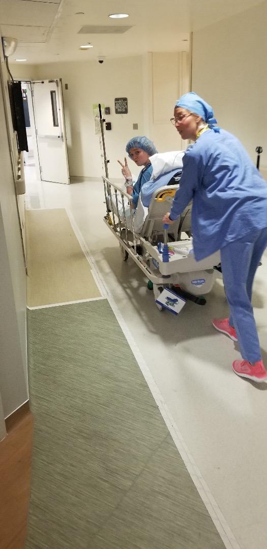 Una mujer joven hace un signo de paz mientras una enfermera asiática vestida de azul la empuja por un pasillo en una cama de hospital.