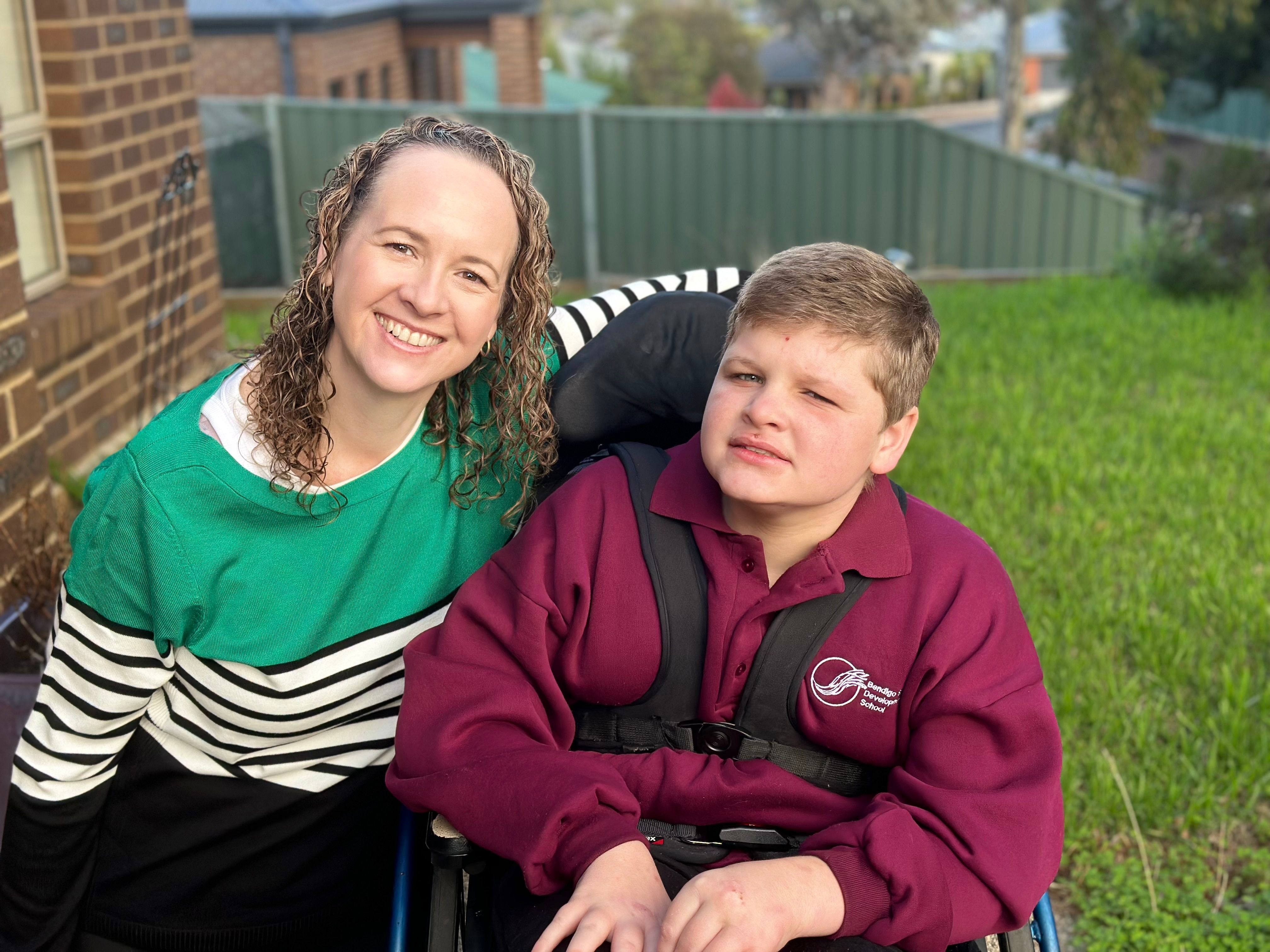 A woman smiling, couched beside a 12 year old boy in a wheelchair.