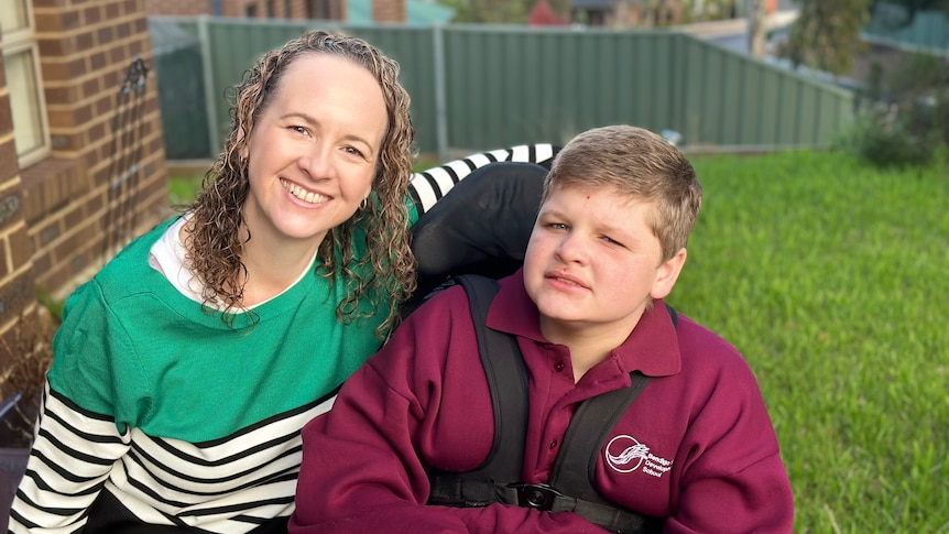 A woman smiling, couched beside a 12 year old boy in a wheelchair.