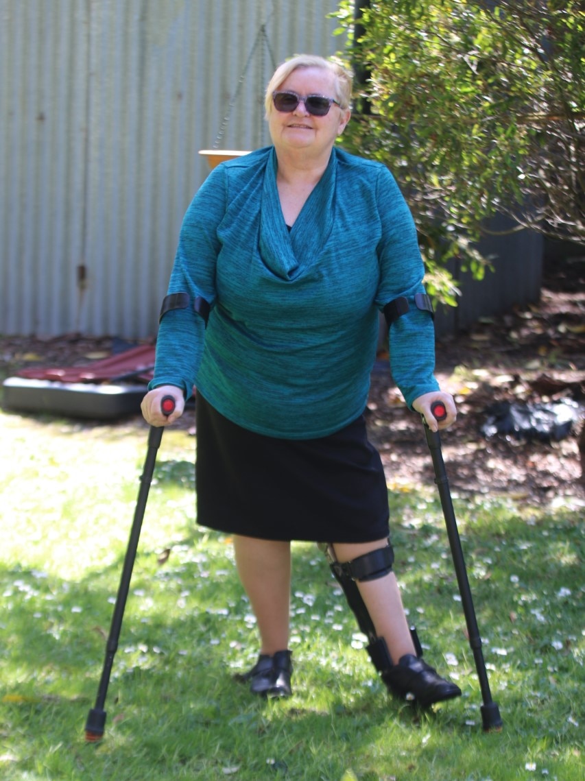 An older woman with blonde hair using crutches. She has sunglasses on and a blue blouse