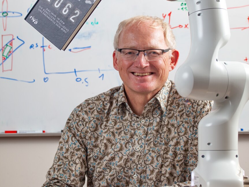 A middle-aged white man with glasses sits at a desk in front of a whiteboard, smiling as a robot holds a book in front of him