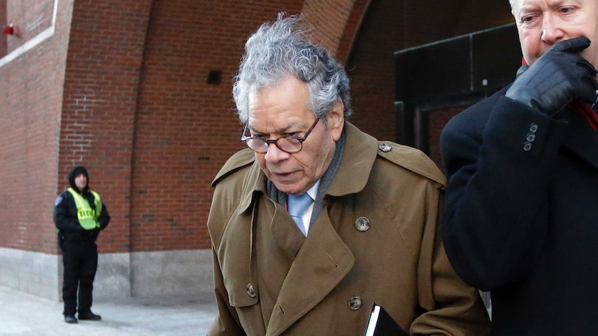 A man wearing glasses and a brown coat puts his head down to avoid cameras outside court.