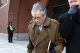 A man wearing glasses and a brown coat puts his head down to avoid cameras outside court.