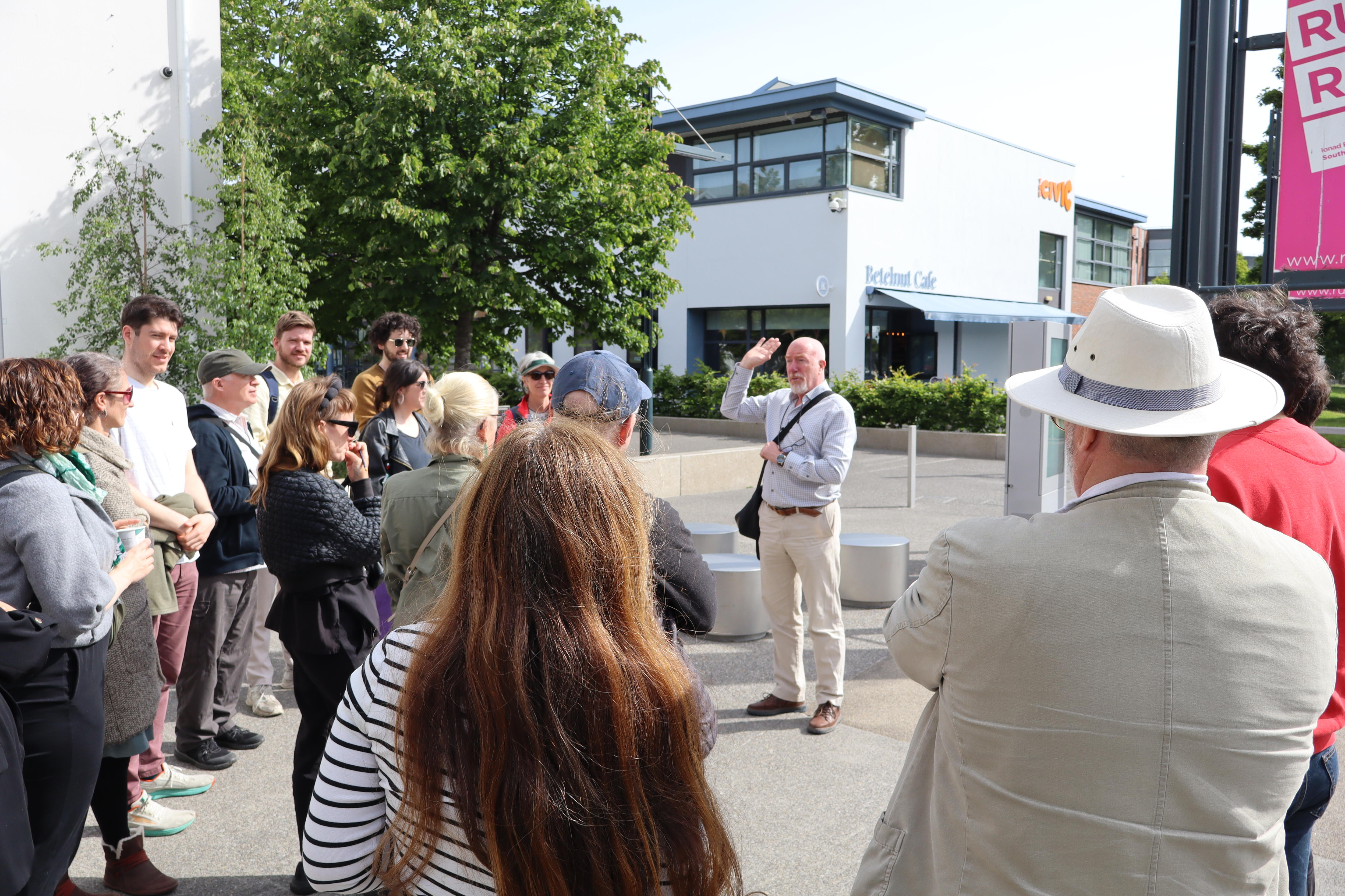 A male tour guide stands surrounded by a small group of people outdoors outside a building. 