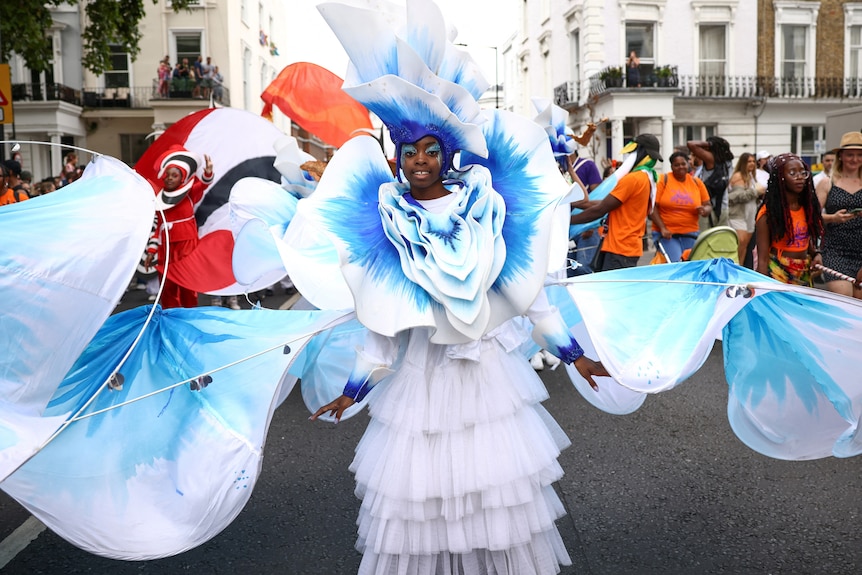 A woman dressed as a white and blue flower twirls her costume on a road during a festival. 