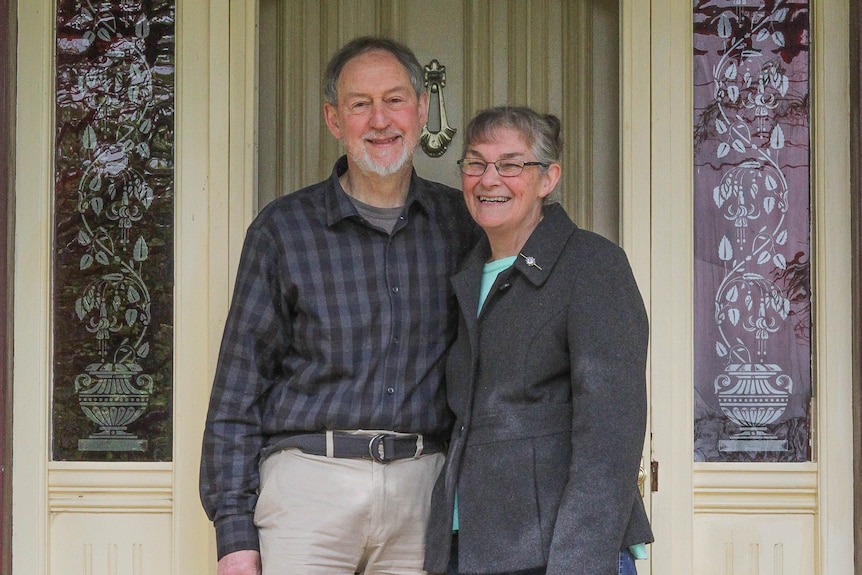 A happy couple of retirement age stand holding each other on the doorstep of a house with an ornate front door